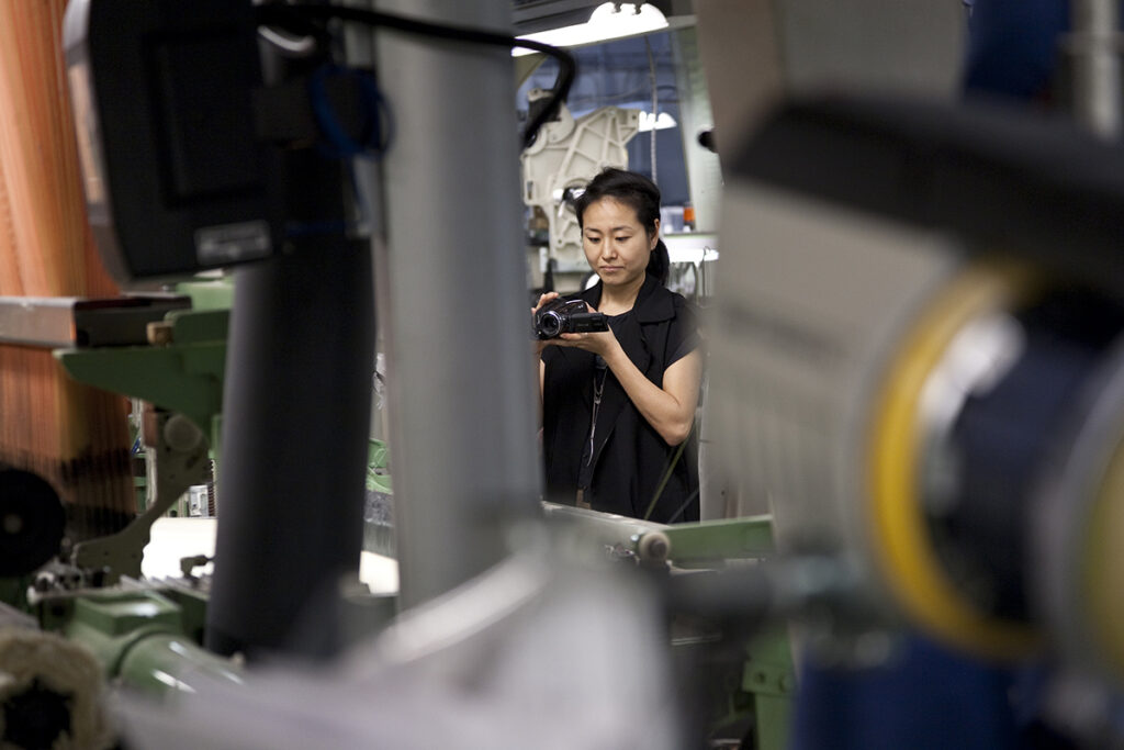 A woman is seen holding a video camera and recording large equipment in an industrial space.