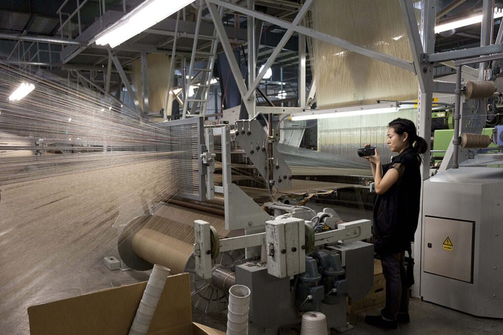 A woman is seen holding a video camera and recording a large Jacquard loom in an industrial space.