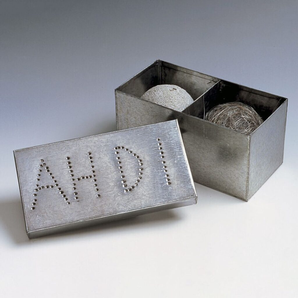 A photo of an opened tin box with the lid leaning against it against a gray background. The tin box is split into two cubes, each with a sphere inside. Inside the left cubicle the sphere appears light gray as if made of cement. Inside the right cubicle, the sphere is brown and appears to be made of a fibrous material. The lid, which faces the viewer, has the initials 