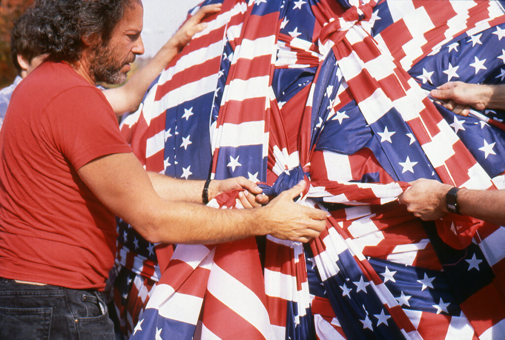 A white man with bushy dark hair and wearing a red shirt works with his hands on an artwork composed of American flags. The flags are being stretched, twisted, and tied by hands pulling in several directions.
