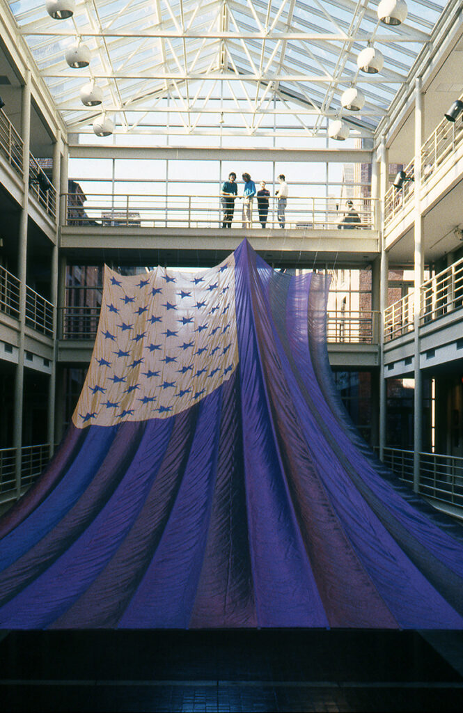 Four people stand, and one crouches on the top mezzanine of a four-storied building. The roof above them is glass, allowing sunlight through. Below them is a giant American Flag pulled by wire to attach to the top mezzanine railing. The slide of the flag closest to the viewer is stretched taut but not yet pulled into position, creating a gentle slope of fabric.
