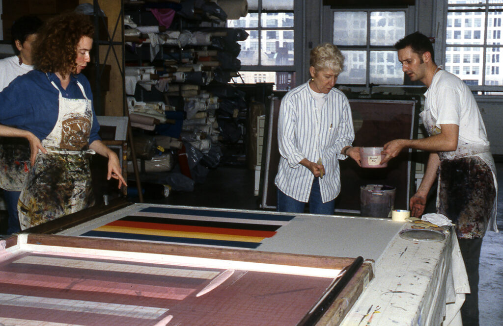 Artists working in a studio. One passes a tub of ink to another artist. They are standing near a large print table with a design composed of colorful stripes.