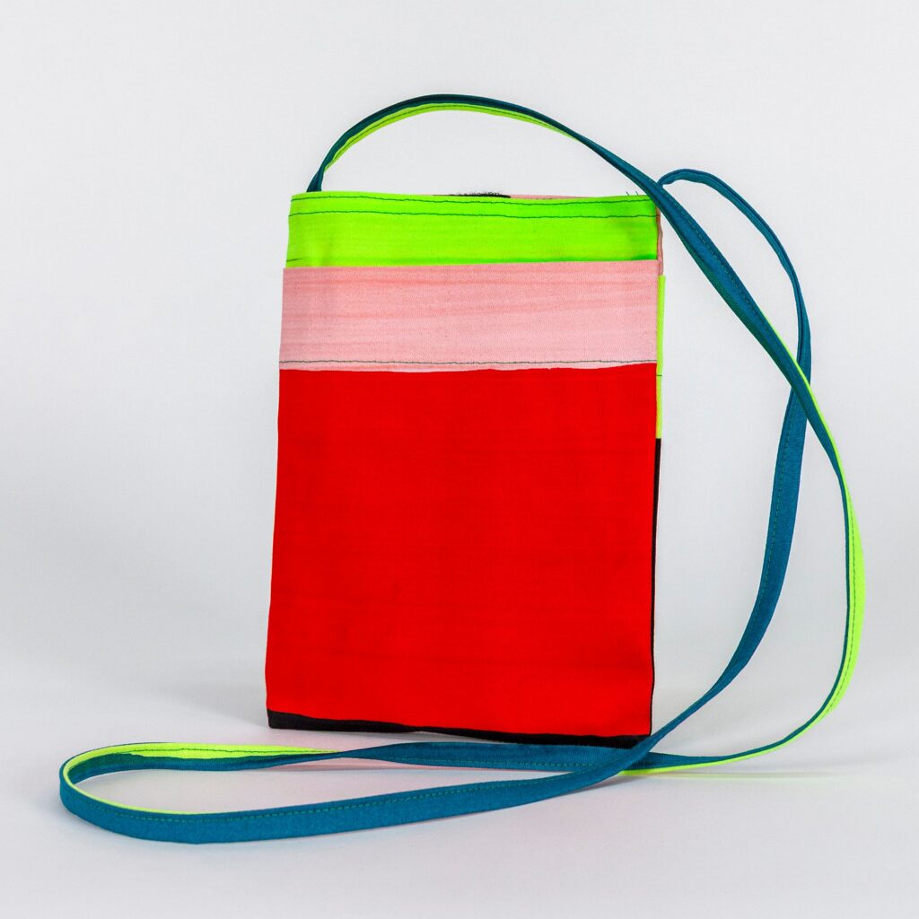 This small, rectangular bag features bold light pink, red, and neon chartreuse stripes. The thin crossbody length strap is neon chartreuse on one side and teal on the other.