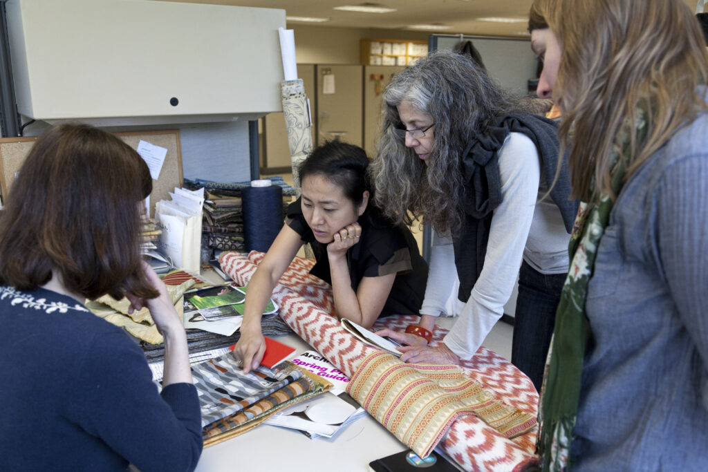A group of women are leaning over a studio table consisting of a roll of fabric and other materials.
