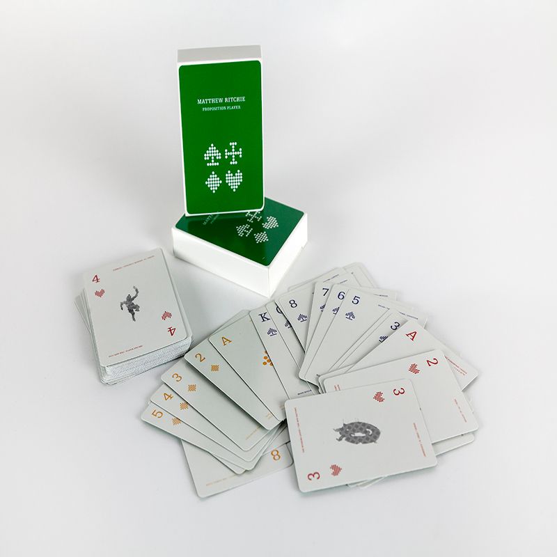 A deck of playing cards sits fanned out on a white backdrop, facing up. In the back, the box they are packaged in is displayed—it is a bright evergreen color with stylized renditions of diamonds, spades, hearts, and clubs in white. At the top, the box is labelled 
