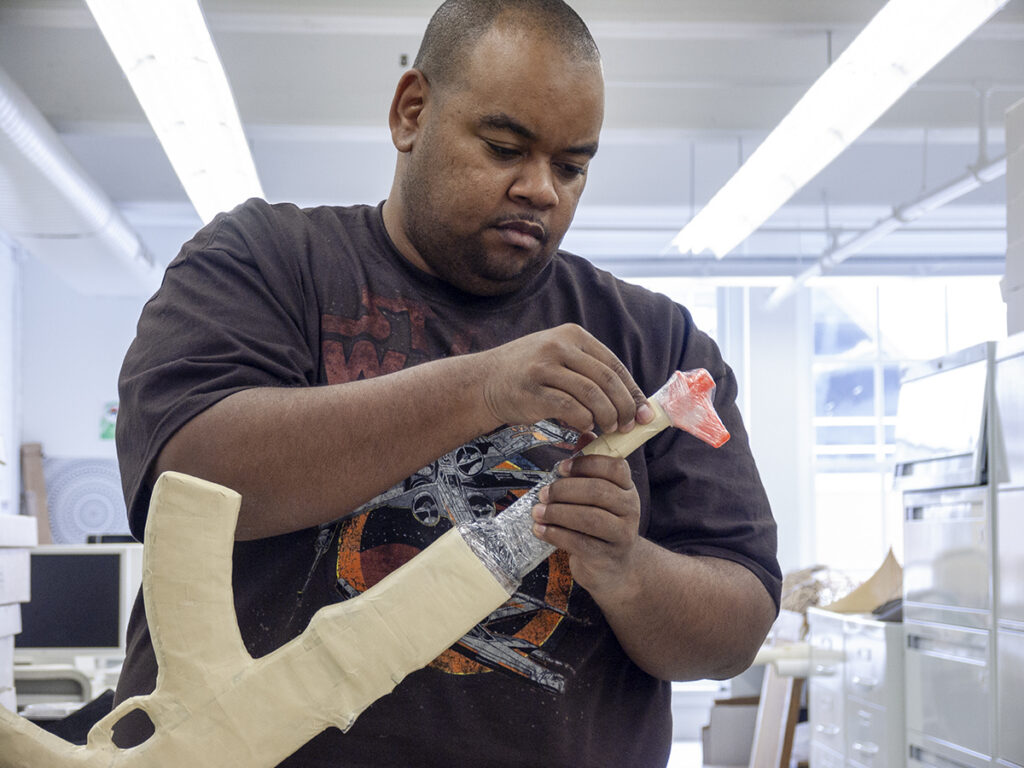 The artist Robert Pruitt, a middle aged Black man, holds a replica automatic rifle while fastening material to its nose
