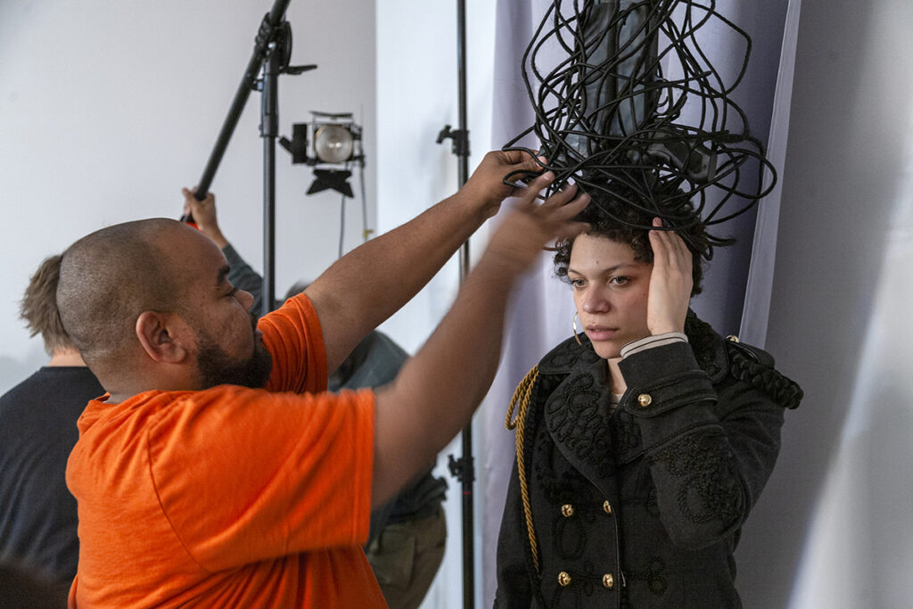 An artist adjusts golden gun necklaces on a model who is seated for a portrait. The model is a Black man wearing a large golden headdress with a partial bicycle wheel inlaid with braids.