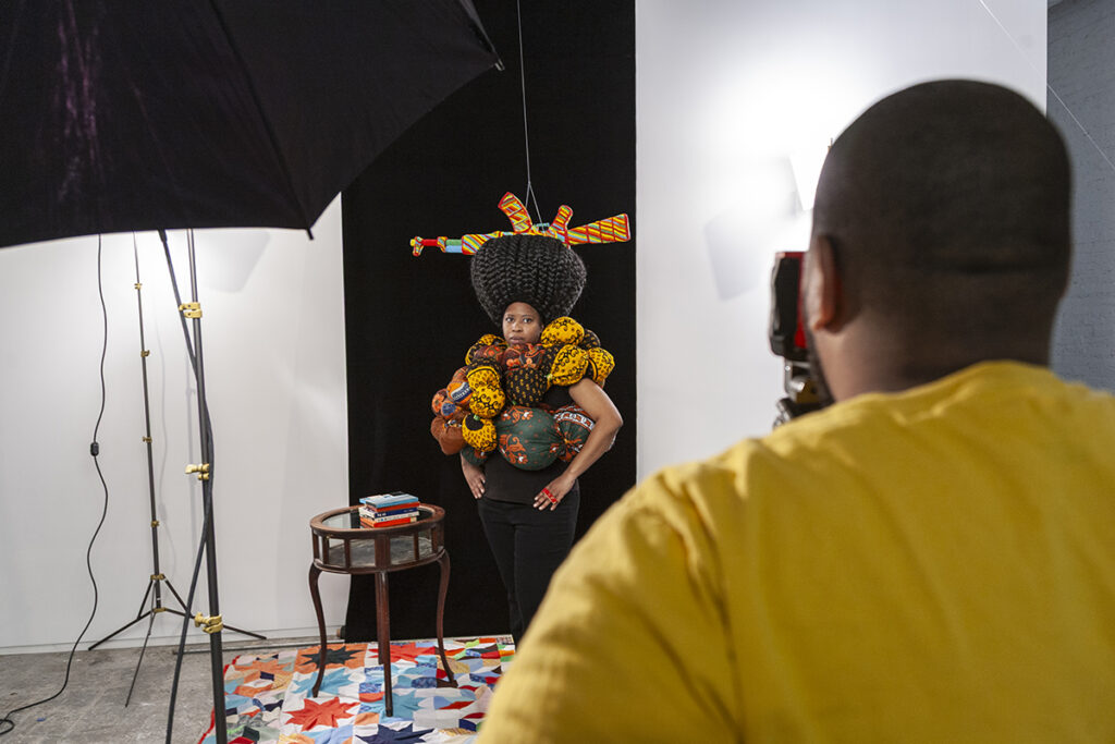 An artist in the foreground points his camera at a model staring back at him. The model is a Black woman wearing bulbous sculptural fabric with patterns on them. A colorful rifle hangs upside down within her large braided hair.