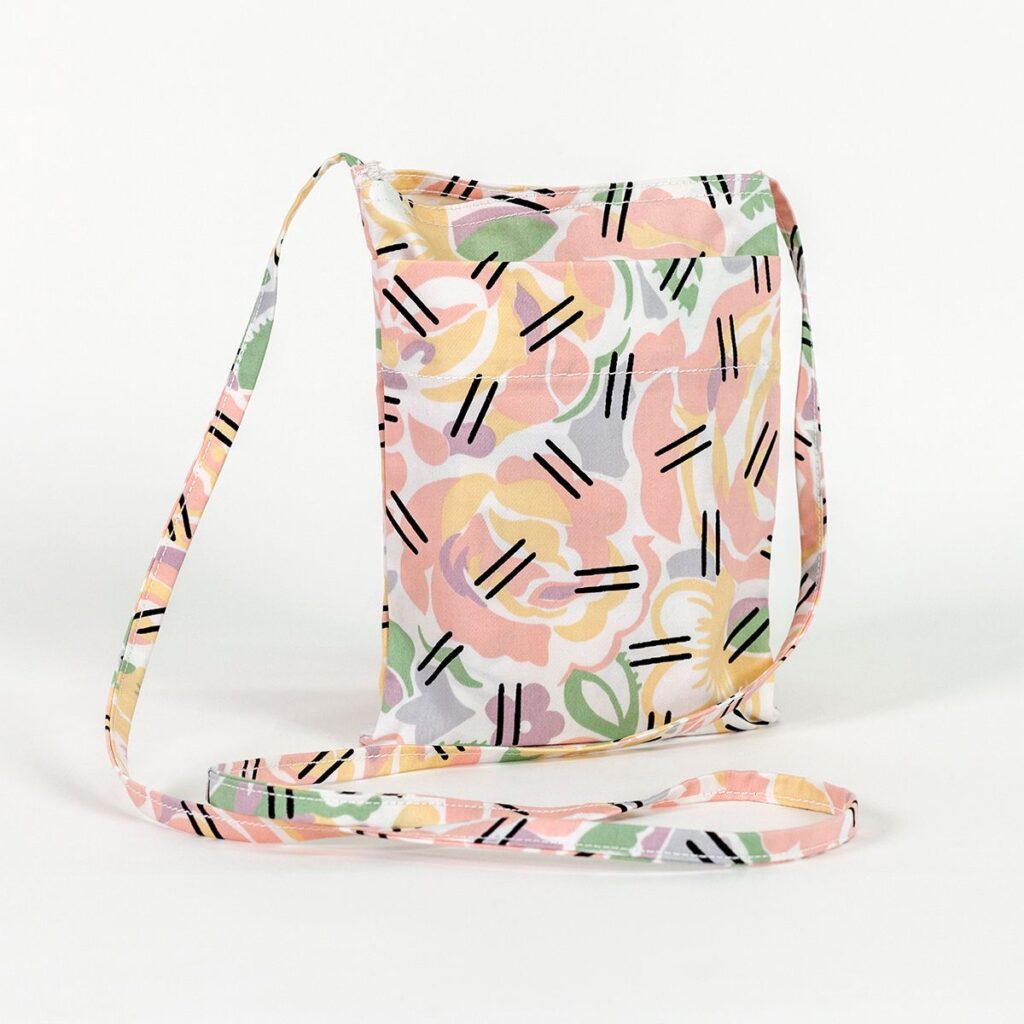 A small flat bag stands upright with long thin straps folding naturally on a surface. The bag features light pastel-colored flowers with overlapping black hatch marks.