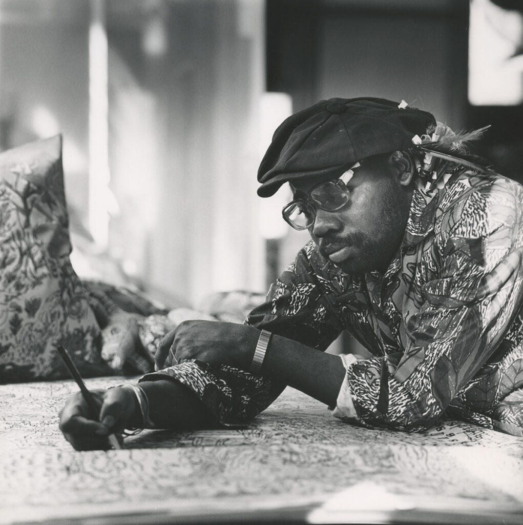 A black and white photograph of Will Stokes, Jr., a Black man with a stylish dress shirt, cap, and watch. He is leaned over a table, drawing a picture.