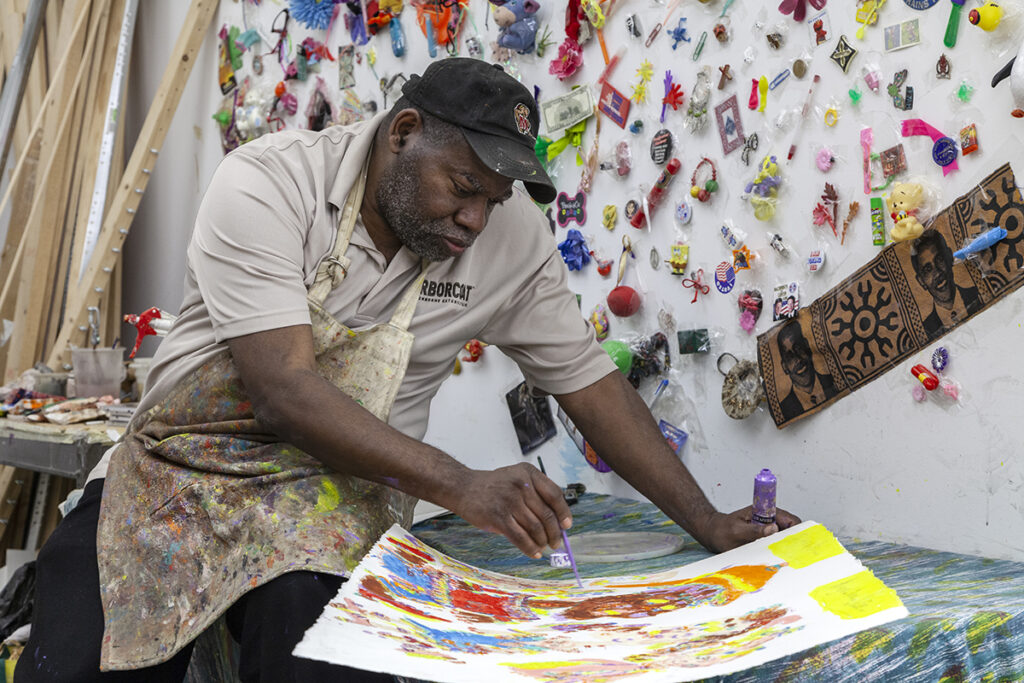 A photo of Will Stokes, Jr., an older Black man in a painter's apron and baseball cap, working on a painting on paper in his studio. He is leaned over a desk working on a warped painting surface. Behind him, many shiny and colorful plastic objects are fixed to the wall.