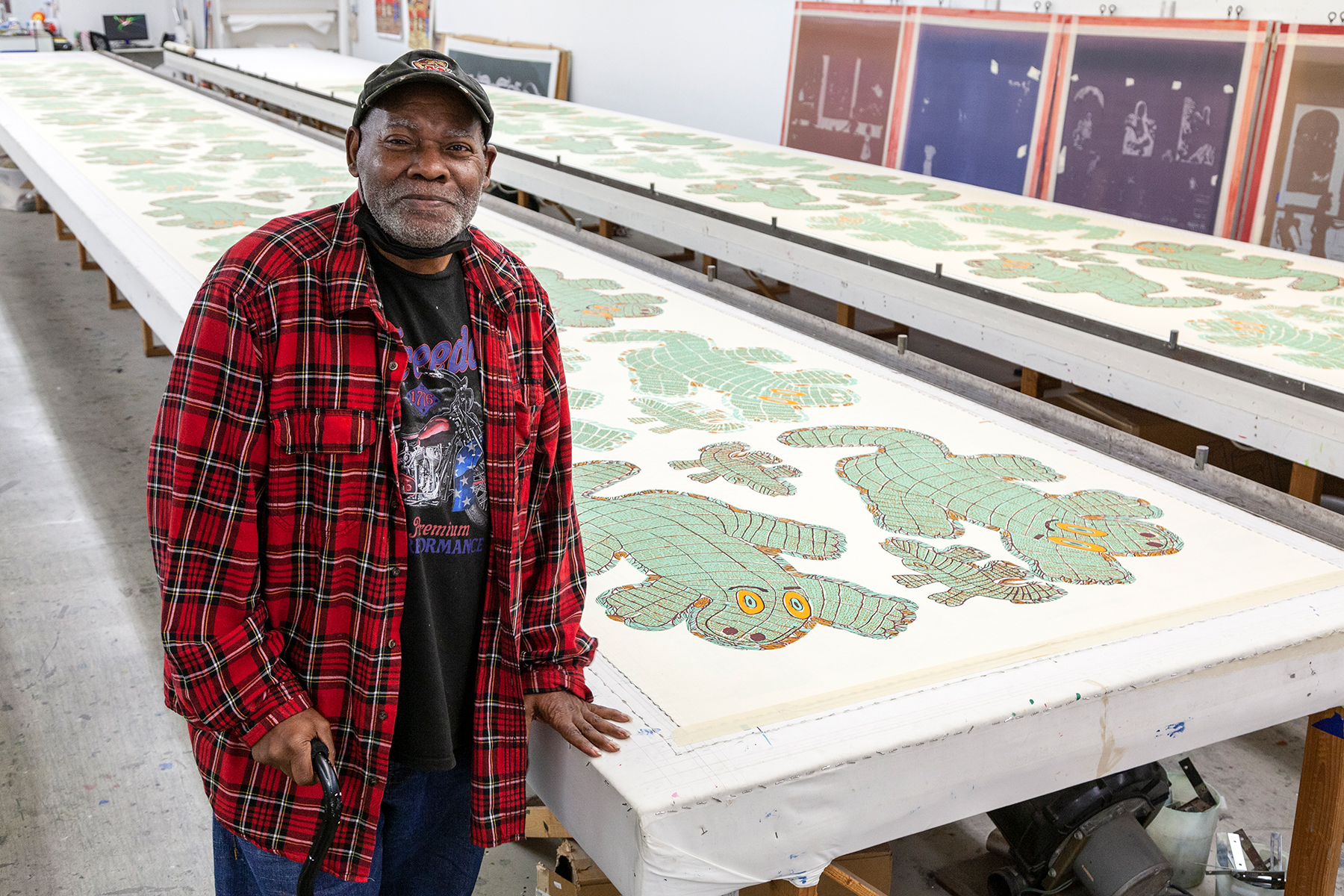 An image of Will Stokes, Jr., an older Black man in a flannel shirt and baseball cap, standing in a print studio. The print tables feature his yardage design of alligators.