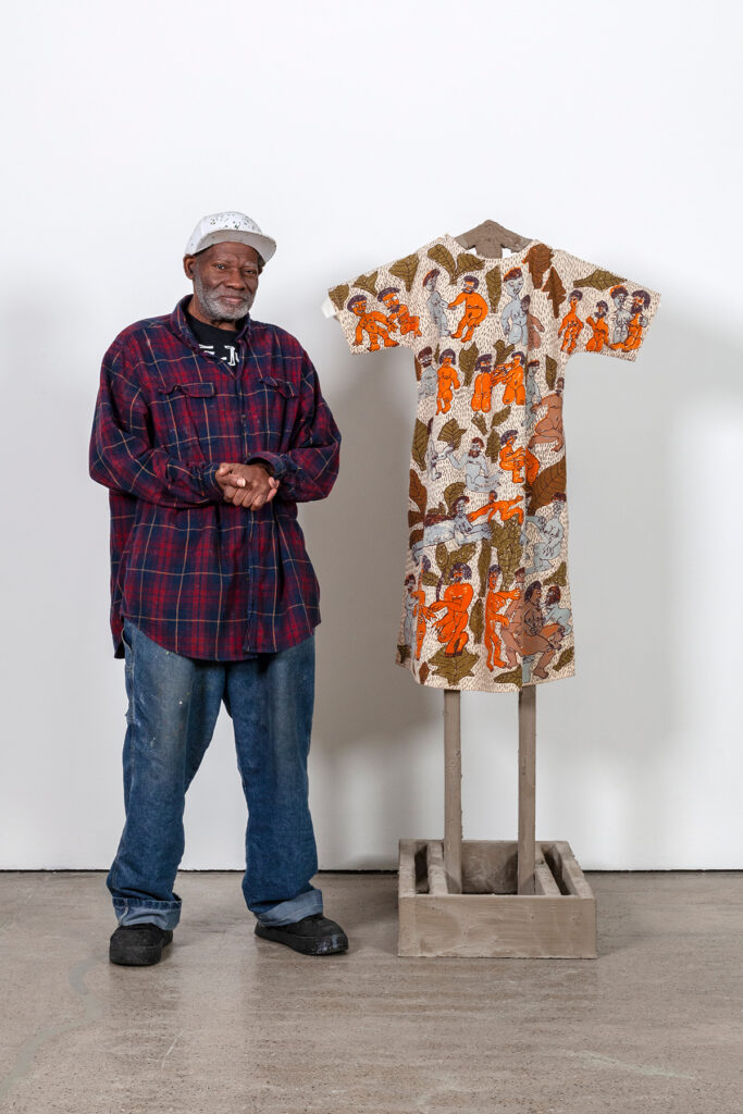 An image of Will Stokes, Jr., an older Black man in a flannel shirt and baseball cap, standing with his artwork in a gallery. His art is a simple dress shape, densely detailed with brown and orange figures, draped over an armature.