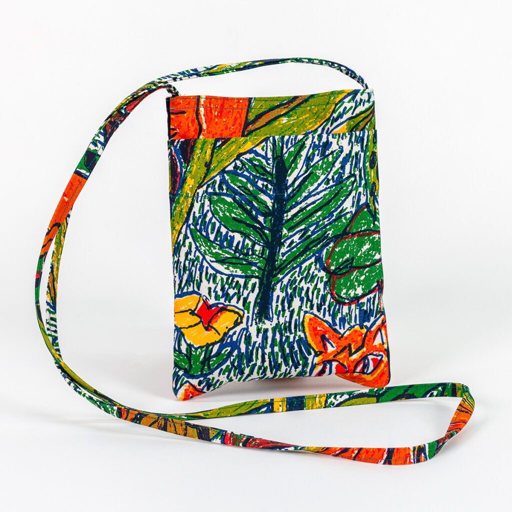 A small flat bag stands upright with long thin straps folding naturally on a surface. The bag features a colorful fabric print with a leafy tree surrounded by hatch markings that suggest grass, a yellow flower, and the eyes of an orange jungle creature.