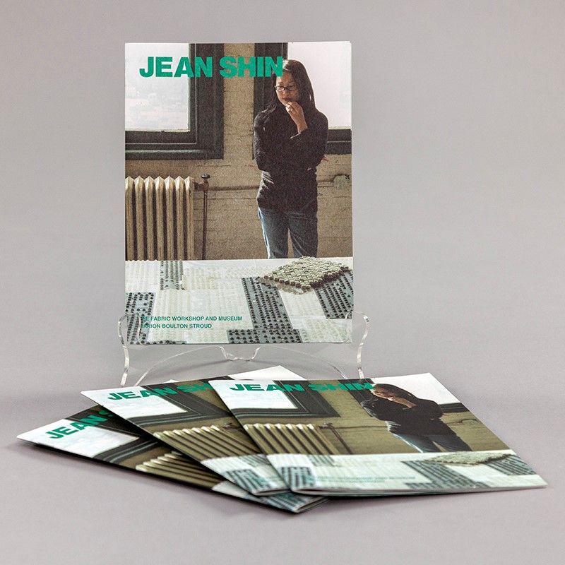 A photo of a book propped up and in the foreground more of the same book spread out as a display. The cover of the book features an artist contemplating her work in progress in a studio. She is looking down at a table where she is working with obsolete keyboards and you can see the underside of several keyboard panels.
