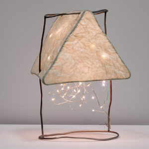 An upright paper lamp glows warmly from within a triangular armature with string lights spilling out below. It is suspended above a table surface from two crooked rods that bridge two circular-shaped rods.