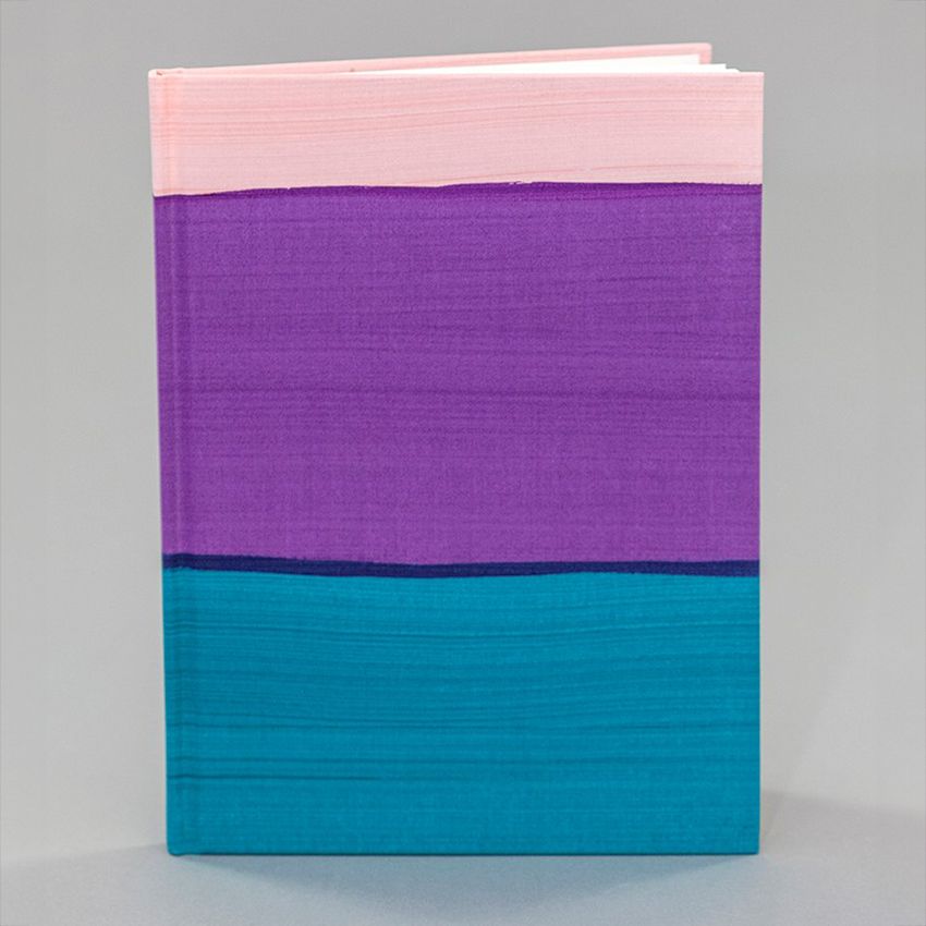 A sketchbook stands upright. Its cover is composed of broad peach, purple, and teal painted stripes.