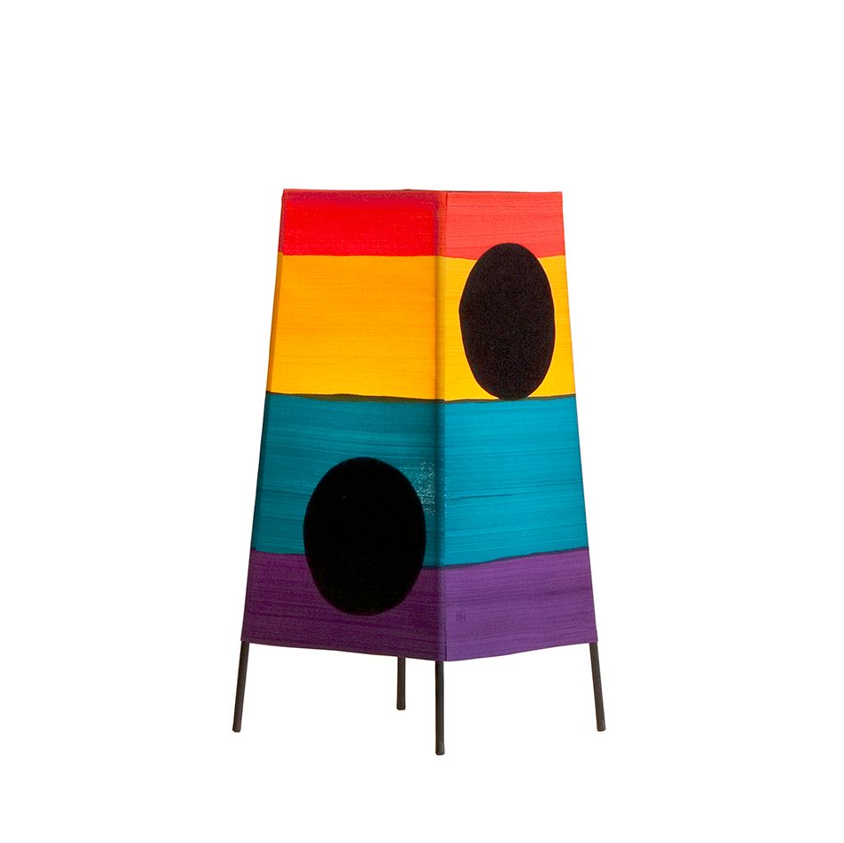 A lamp sits against a white background. It has four black metal thin legs arranged in a square which go up, and are then wrapped in fabric. The fabric has a horizontal purple stripe at the bottom, a teal stripe above the purple, a yellow stripe above the teal, a red stripe at the top, and two large black dots—one on each side of the lamp visible to the camera.