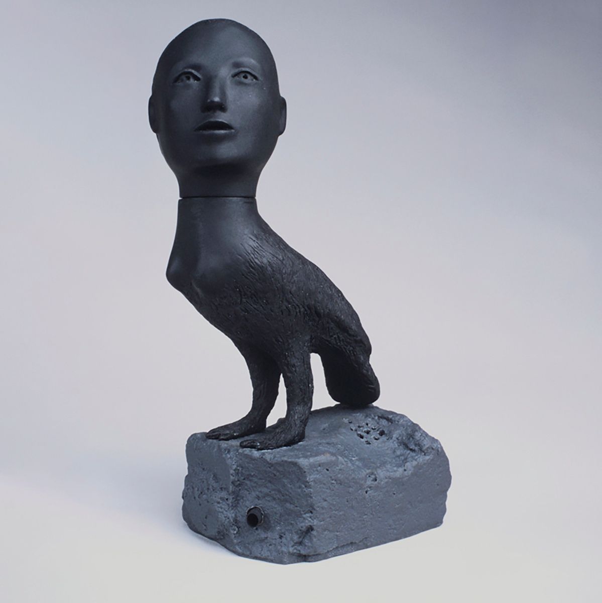 A small sculpture of a creature composed of a human head and a bird's body perched on a small rock.