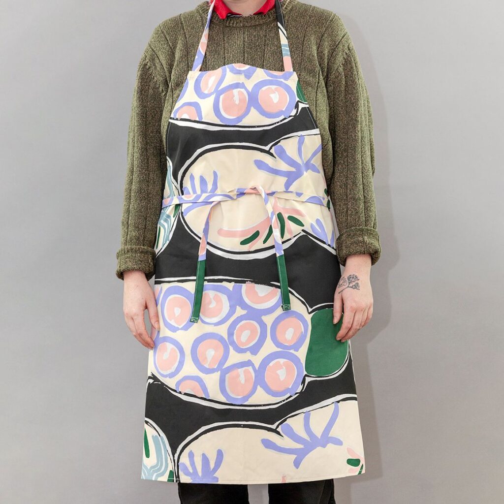 A photo of a person modeling a cooking apron. The apron is a screenprinted design by Betty Woodman. The background of the design is black, with colorful pastel, loosely drawn, soft abstract shapes mimicking ceramic platters.