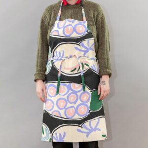 A photo of a person modeling a cooking apron. The apron is a screenprinted design by Betty Woodman. The background of the design is black, with colorful pastel, loosely drawn, soft abstract shapes mimicking ceramic platters.