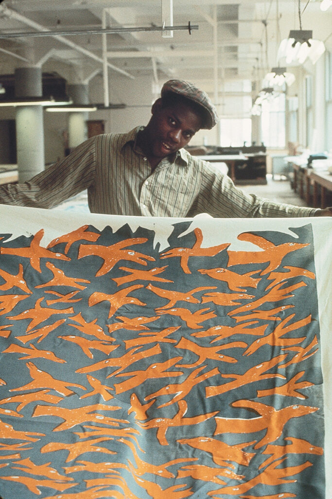 An image of the artist Will Stokes, Jr. as a young man. Will, a Black man in a dress shirt and cap, is spreading out his yardage design featuring orange birds flying in all directions over a blue ground.