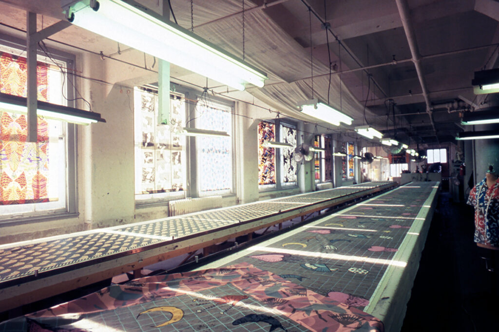 A large artist studio with very long printmaking tables. A design featuring hearts, stars, fish, dice, hands, and the moon is depicted repeatedly along the tables. There is another identical table next to it with a different design, and several windows on the wall with sheer printed fabric overtop.