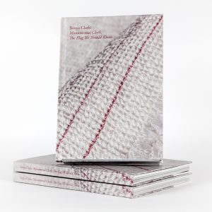 A photograph of a book propped upright on top of a pair of laterally stacked copies of the same book. The cover of the book features a cream-colored woven textile with two thin red stripes that stretch from corner to corner. The text on the top-left corner reads, 