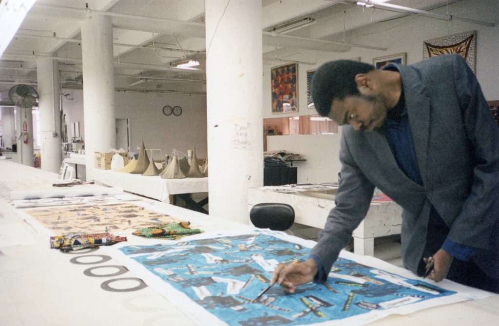 Color photograph in landscape orientation. Photograph is a process documentation of the artist, Yinka Shonibare, designing his fabric yardage titled “South Street”. The artist is a Black man wearing a blue shirt under a grey suit jacket along with black pants. He is holding an object in each hand as he renders the design for his piece. The print on the table in front of him is blue with words printed over it in yellow, black, and white. On the rest of the table, there are additional materials that have prints on them. In the background, there are other pieces either hanging on a wall or sat on a table.