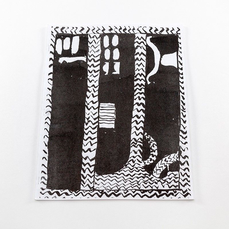 An image of a tall book laying flat on a surface. The cover is regular, white paper. Printed on the white paper is an image of a drawing illustrated in black ink. The drawing features three black columns. In the space between the columns is a gestural zig-zag pattern. There are also small, organic shapes towards the tops of the column with each column having its own unique set of shapes.
