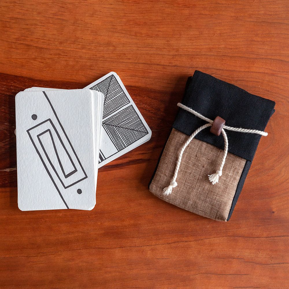 The image shows a sewn fabric pouch in black and brown linen with a white rope and dark brown clay bead closure. To the left of the pouch is a stack of cards letter pressed in black ink on a cream paper, with abstract designs. Both items are against a wooden background.