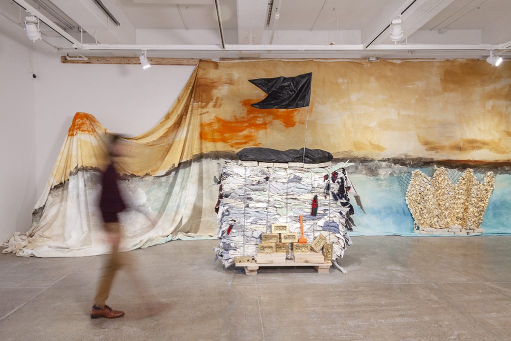 A contemporary art installation by the artist Henry Taylor. A person walks near a totem created from a bale of house siding gathered as post-industrial waste. Atop the totem is a black flag. Behind it is a large painted backdrop, draping from the ceiling.