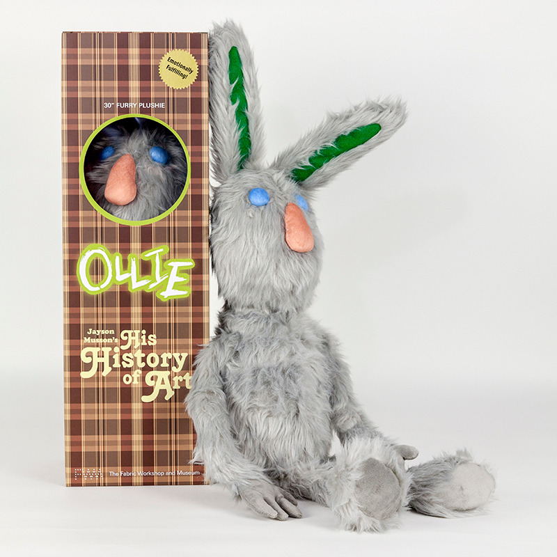 A stuffed plush gray rabbit sits upright against a plaid collector's box. The rabbit has round blue eyes, an oblong peach nose and long green shapes on the interior of its ears. The brown plaid box reads 