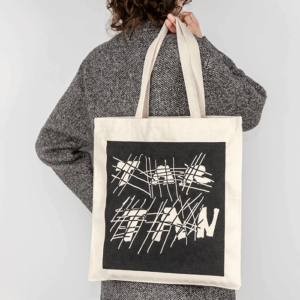 A person wearing a grey coat is facing away from the camera against a white backdrop. Slung over their shoulder in their right hand and facing the viewer, is a cream colored canvas tote with a black ink square screenprinted over the front of the bag. The negative space in the print has crosshatched lines intersecting with thick handwritten text that reads 