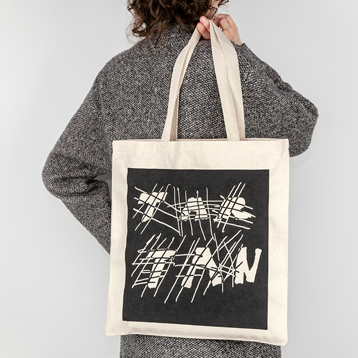 A person wearing a grey coat is facing away from the camera against a white backdrop. Slung over their shoulder in their right hand and facing the viewer, is a cream colored canvas tote with a black ink square screenprinted over the front of the bag. The negative space in the print has crosshatched lines intersecting with thick handwritten text that reads 