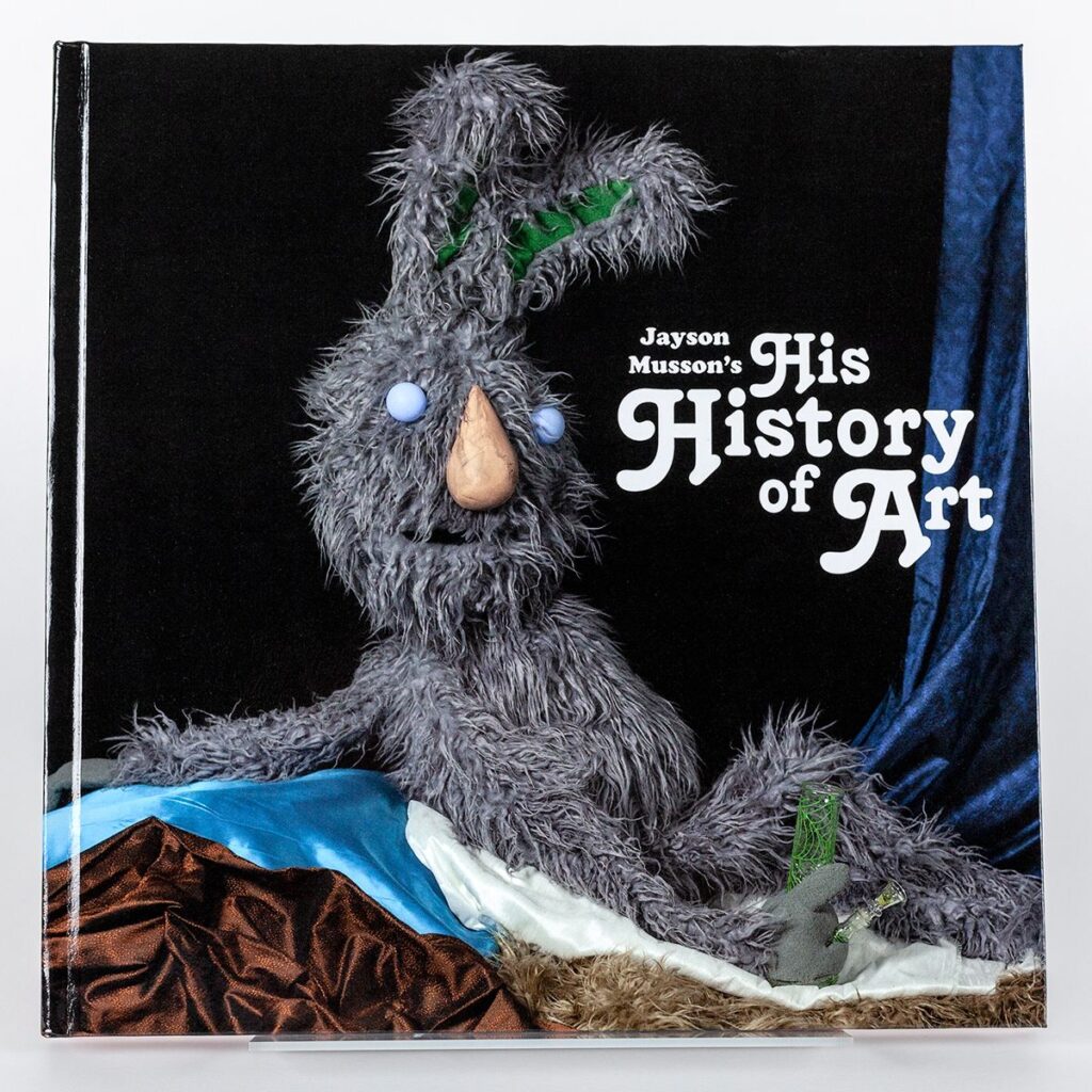 A cover of a book featuring a gray scraggly rabbit as a puppet lounging in reference to Ingres' painting 