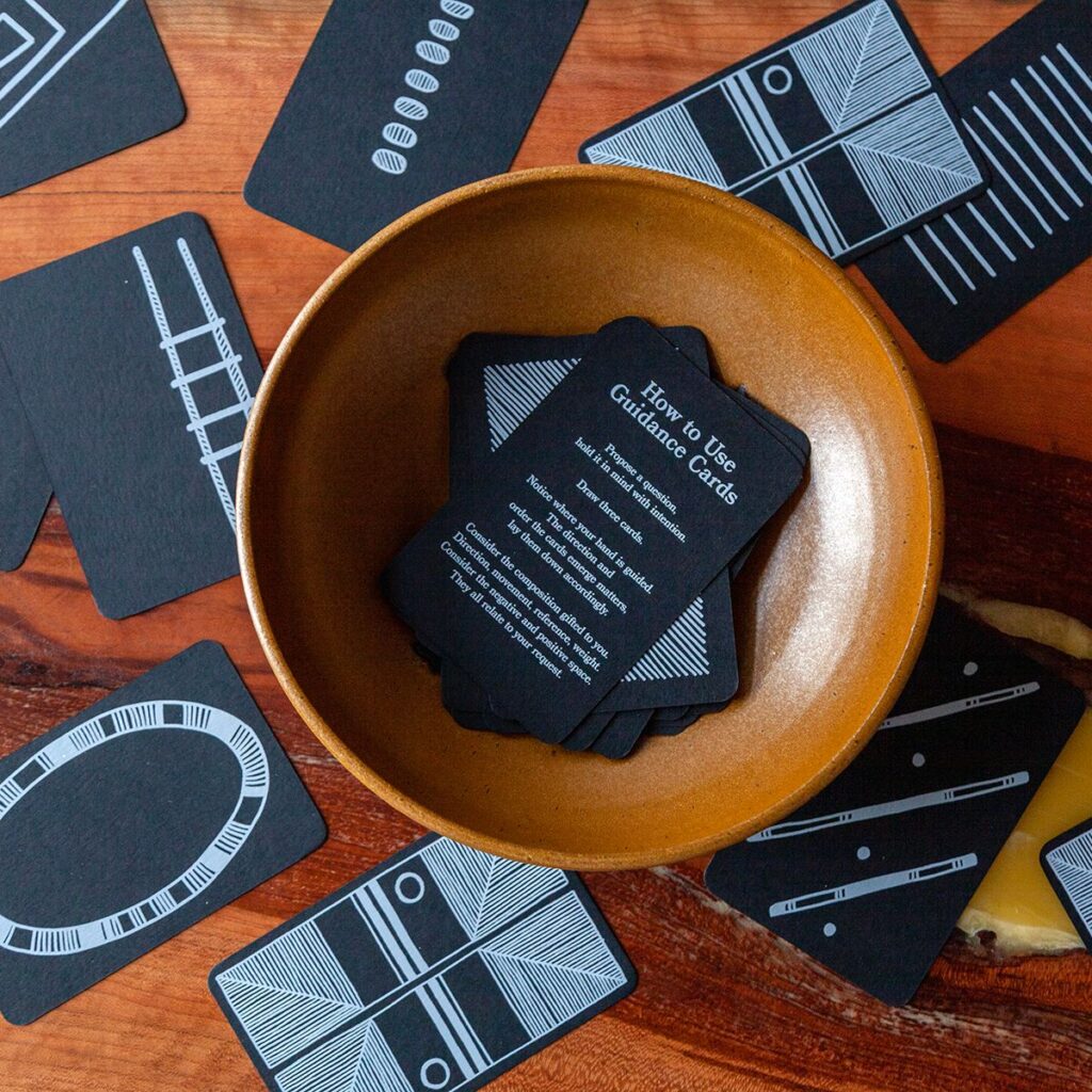 A bird's eye view of a rich, earth toned ceramic bowl sitting on a wooden tabletop. Inside the bowl is a stack of cards, the top card is instructional and reads 