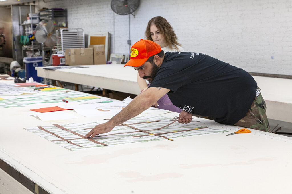 A photo of the artist Borna Sammak, a brown-skinned man with a dark beard, as he leans over the table to arrange drawings on a long table. A woman with curly hair watches as he works.