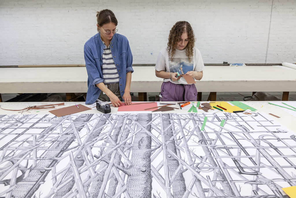 A photo of two women studio artists cutting and arranging scraps of paper. They are standing in front of a long black and white repeat pattern of trees and branches.