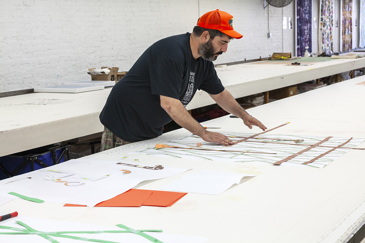 The artist Borna Sammak, a brown-skinned man with a beard, lays out drawings on a long table. He is wearing a bright orange hat and a black t-shirt.