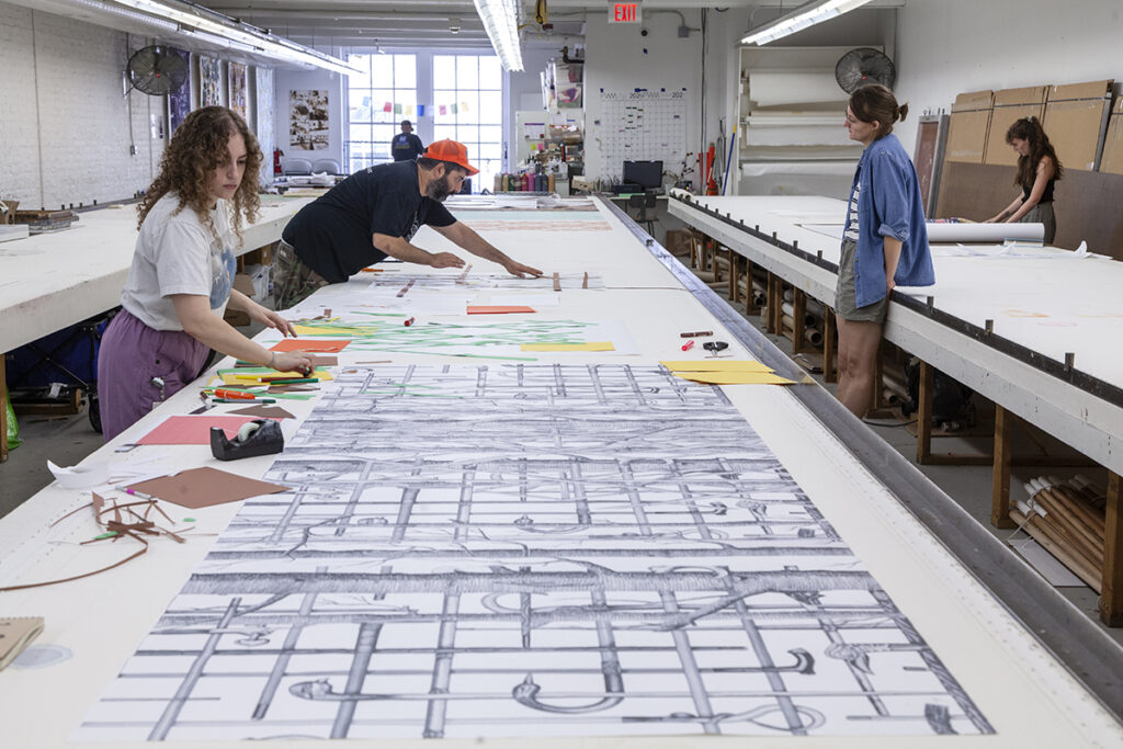 A photo of three artists working from various sides of a long table. They are arranging drawings and scraps of paper over a long repeat pattern of gridded lines.