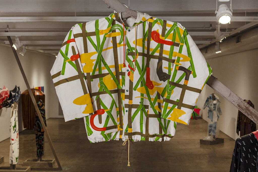 A photo of a jacket hanging in a gallery. It features various intersecting lines in green, brown, and yellow and hints of red.