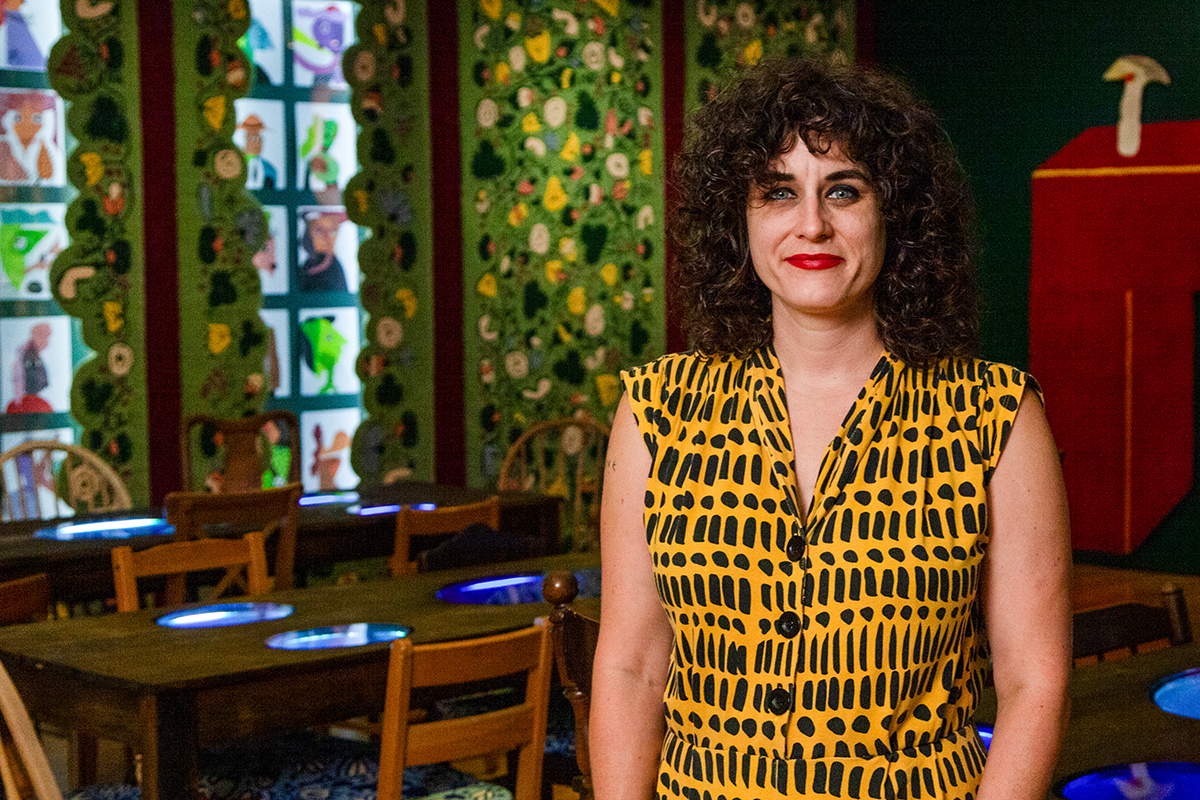 A portrait of the artist Jessica Campbell, a white woman with curly brown hair. She poses for the camera with her tufted floral artwork in the background. She is wearing a sleeveless yellow and black patterned shirt.