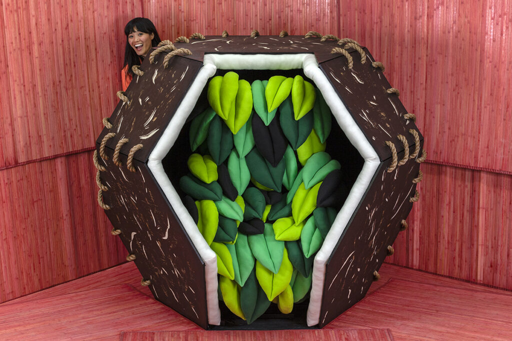 A photograph of the artist Risa Puno, a young woman with black hair, peeks out from behind a brown geometric pod. The interior of the pod contains a seat made of green felted leaves. The walls and floors surrounding them are made of pink bamboo.