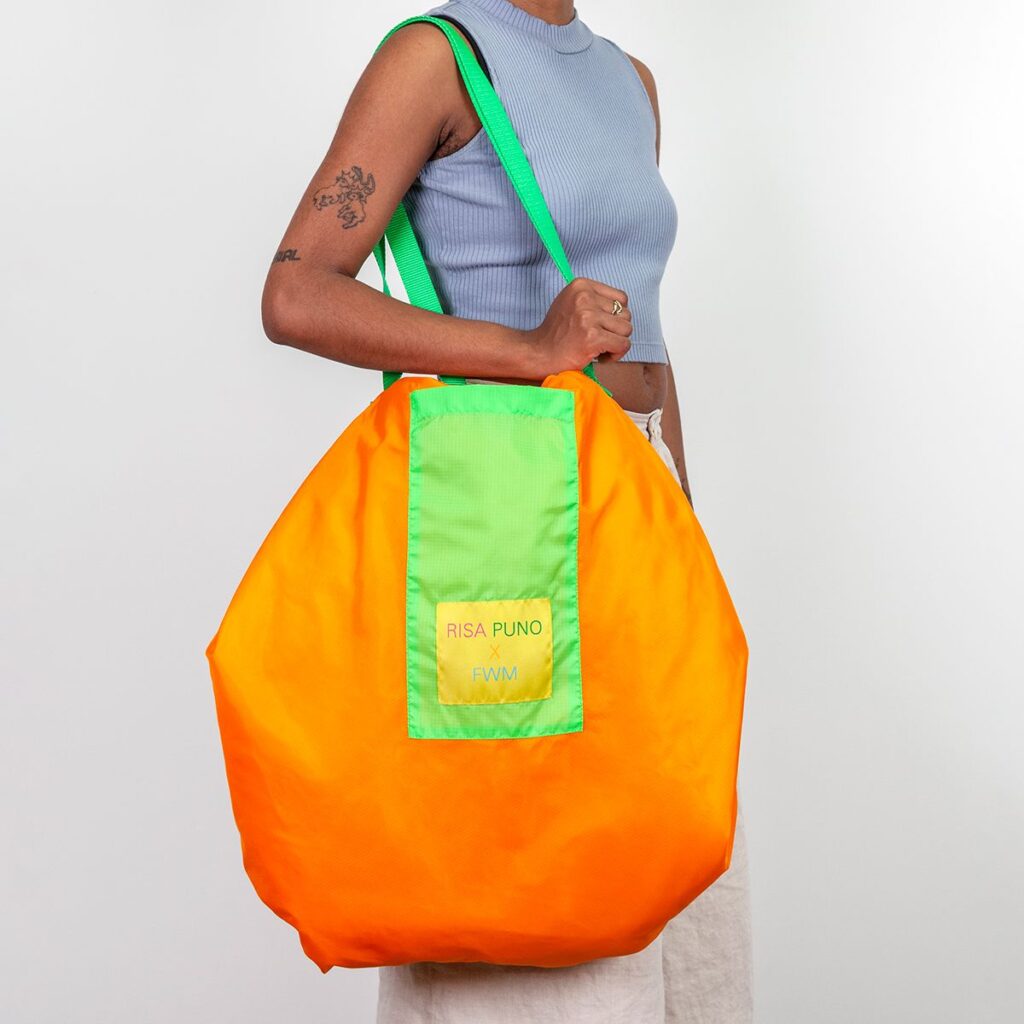 A person holds a large nylon bag on their shoulder. The bag is orange with green straps and a yellow tag that reads 