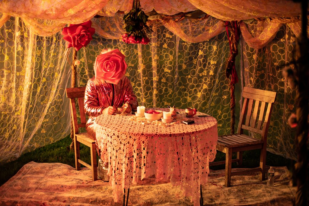 A photograph of a person seated at a round dining table. They are wearing a shiny pink outfit and their face is obscured by a large rose. The table is covered by a draping tablecloth made up of doily patterns. The environment is likewise draped in sheer fabric curtains. There is an empty chair to the right of the table and a bouquet of roses hanging upside down from the ceiling, just over the seated person.