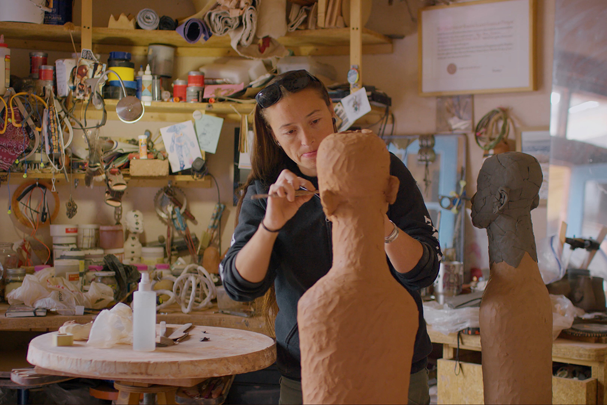 The artist Rose B. Simpson carves two clay figures with hand tools. Behind her are many tools and supplies gathered along shelves in her studio. The image is a still from the Art21 video feature Simpson and other contemporary artists.