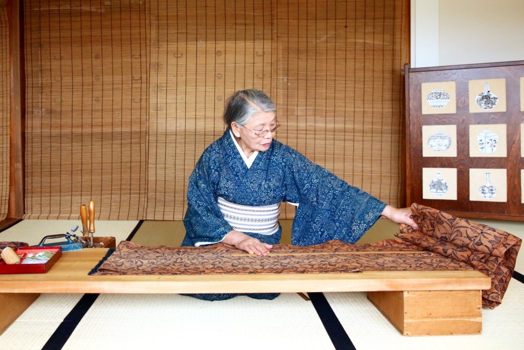 Master Kimono maker Tsuyo Onodera from Sendai, Japan. Picure of an older Japanese woman seated at a low table unfurling a scroll of fabric.