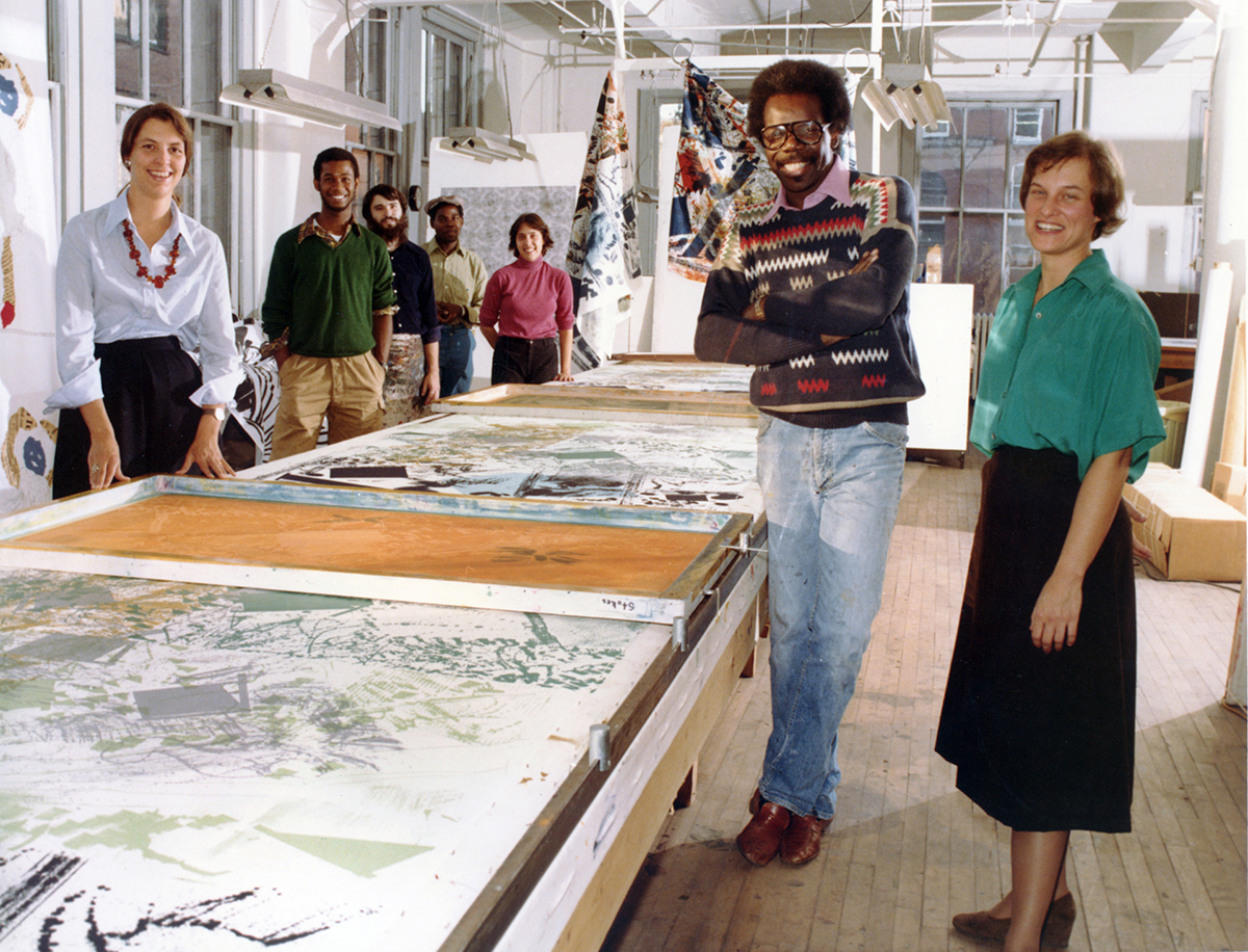 Several people gather around a large printmaking table. To the right is the artist, a tall Black man crossing his arms, smiling. At his right is a shorter white woman, Kippy Stroud, founder of The Fabric Workshop and Museum.