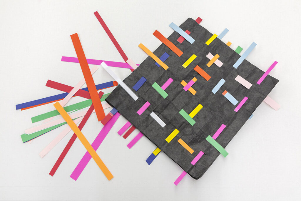 A black square of paper with many strands of thin colorful paper woven through it.